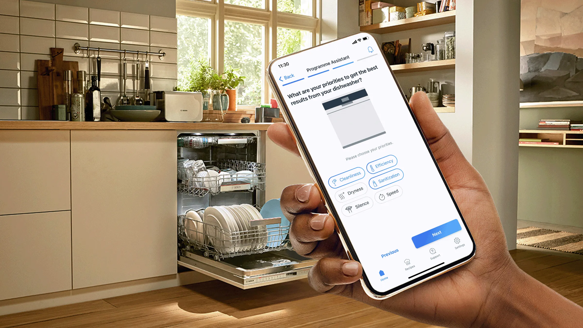 Eine Hand hält ein Smartphone mit dem Home Connect Programm-Assistenten in einer hellen Küche vor einem geöffneten, beladenen Geschirrspüler.