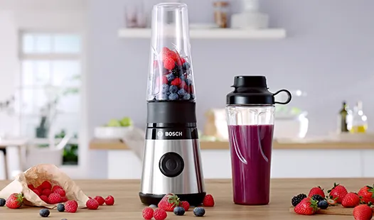 Ein Bosch-Mixer mit Beeren darin, neben einem lila Smoothie in einem To-go-Becher, auf einer Küchenarbeitsplatte mit verstreuten frischen Beeren.