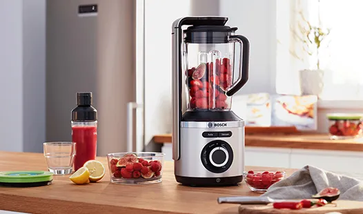„Ein stilvoller silberner Mixer auf einer Arbeitsplatte aus Holz, gefüllt mit Erdbeeren, daneben eine Smoothieflasche, eine aufgeschnittene Zitrone und frische Himbeeren.