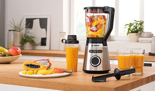 Ein moderner Mixer auf einer Küchenarbeitsplatte, gefüllt mit bunten Früchten, daneben Smoothiegläser und eine Schale mit frischem Obst.