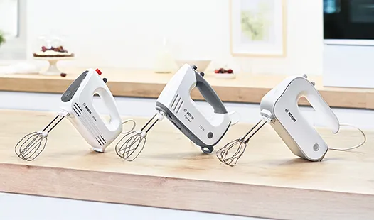 Drei elektrische Handmixer in Weiß und Grau stehen in einer Reihe auf einer hölzernen Küchenarbeitsplatte vor einem hellen, minimalistischen Hintergrund.