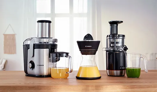„Verschiedene Entsafter in Schwarz und Silber auf einer Arbeitsplatte aus Holz, mit orangem und grünem Saft in durchsichtigen Behältern in einer hellen Küche.