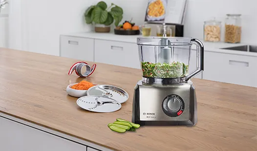 En matberedare i rostfritt stål på en köksbänk i trä, med hackade grönsaker, rivjärnstillbehör och en skål med morötter i bakgrunden.