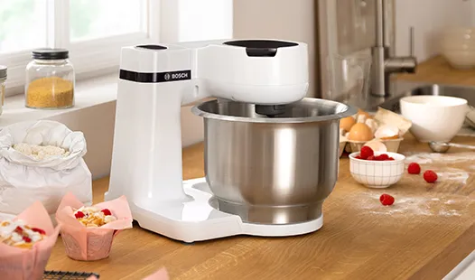 Ein weißer Standmixer mit einer Edelstahlschüssel steht auf einer Holzarbeitsplatte, umgeben von Backutensilien und Zutaten.