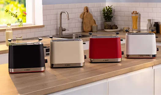 Vier stilvolle Toaster in Schwarz, Beige, Rot und Weiß stehen nebeneinander auf einer Küchenarbeitsplatte, während natürliches Licht hereinströmt.