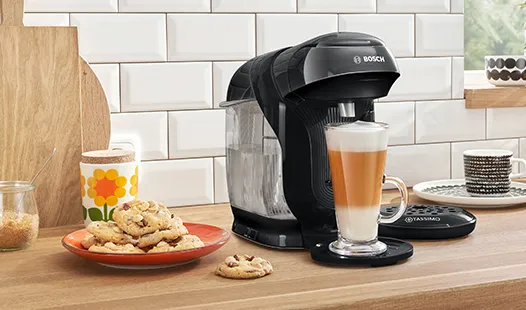 Eine schwarze Kaffeemaschine bereitet einen Latte in einer Glastasse auf einer Arbeitsplatte zu, umgeben von Keksen und bunten Tassen.