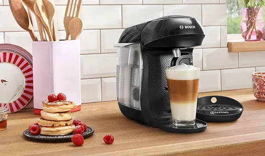 Eine Bosch-Kaffeemaschine steht neben einem Glas mit geschichtetem Latte, Pfannkuchen, die mit Himbeeren belegt sind, und Küchenutensilien in einer gemütlichen Atmosphäre.