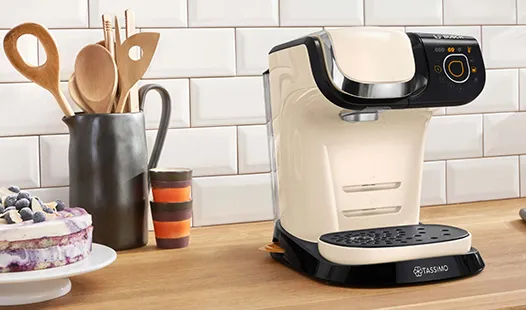 Eine cremefarbene Kaffeemaschine steht auf einer Holzarbeitsplatte, neben Besteck, bunten Tassen und einem Kuchen mit Beeren.