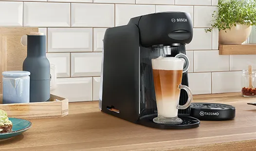 „Eine elegante schwarze Kaffeemaschine bereitet einen Latte in einer klaren Glastasse zu, die auf einer Holzarbeitsplatte steht, mit Küchendekoration im Hintergrund.