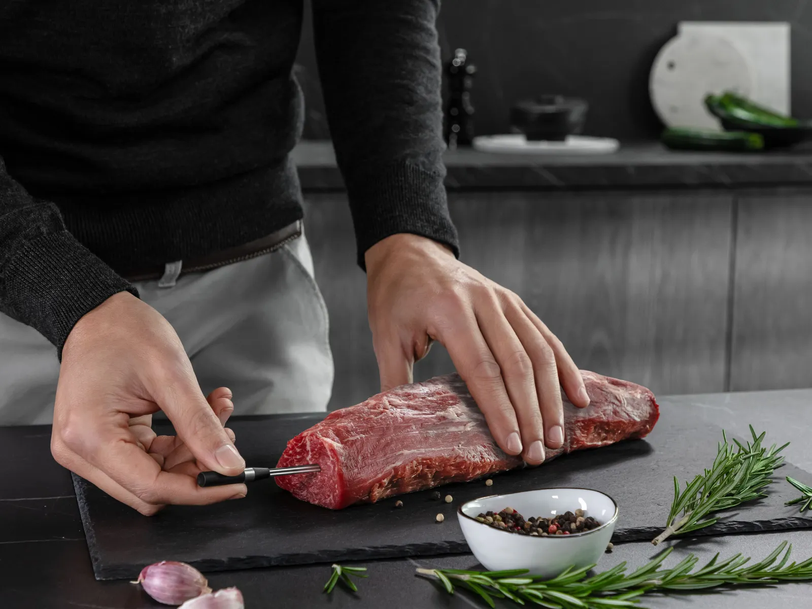 Ein Mann setzt den kabellosen roastingSensor in ein rohes Rinderfilet ein, daneben Knoblauch und Kräuter.
