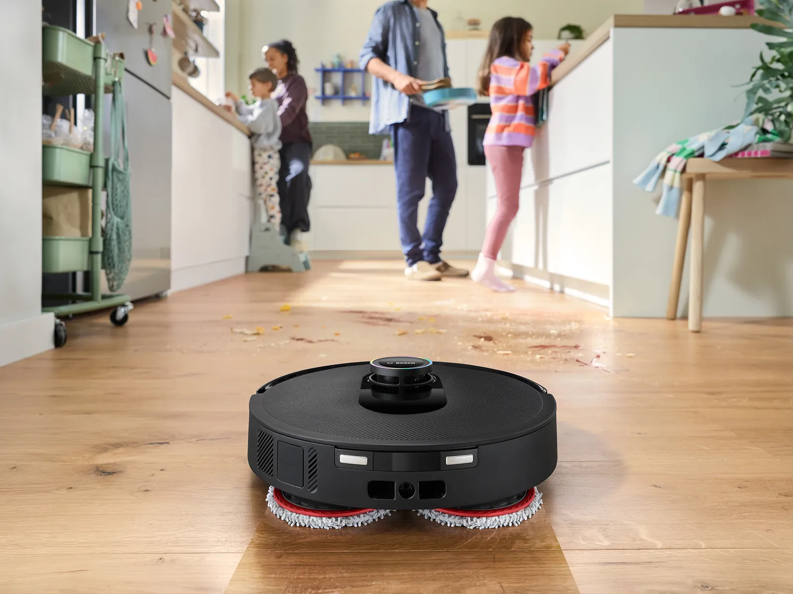 Spotless städrobot som är i gång med att mopa ett trägolv i ett ljust rum med familj i bakgrunden.