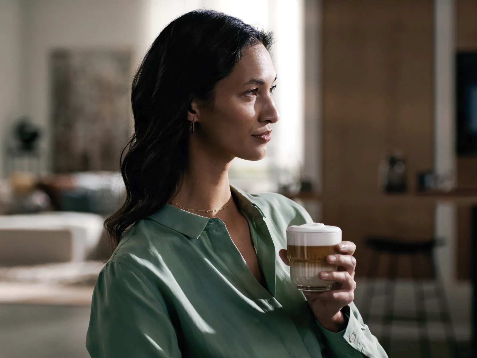 Femeie într-o bluză verde, ținând o ceașcă de cafea într-o cameră modernă.