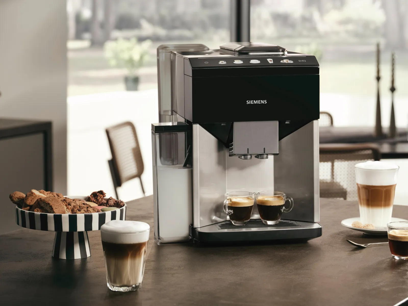 Siemens Kaffeevollautomat auf der Arbeitsplatte, daneben drei gefüllte Tassen und ein Teller mit Gebäck.