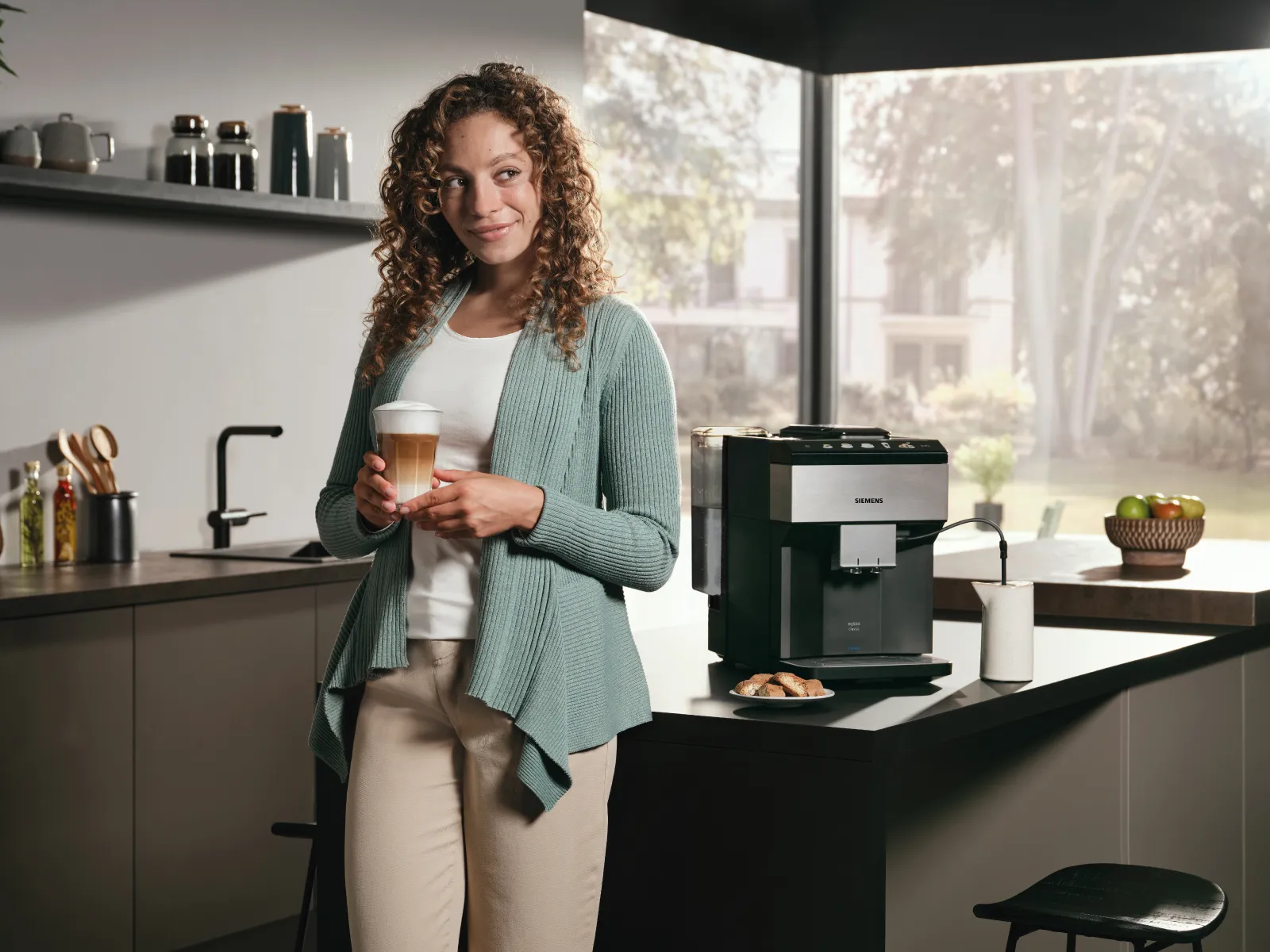 Eine lächelnde Frau mit einem Latte Macchiato in den Händen steht vor einer Kücheninsel; neben ihr steht der EQ500.