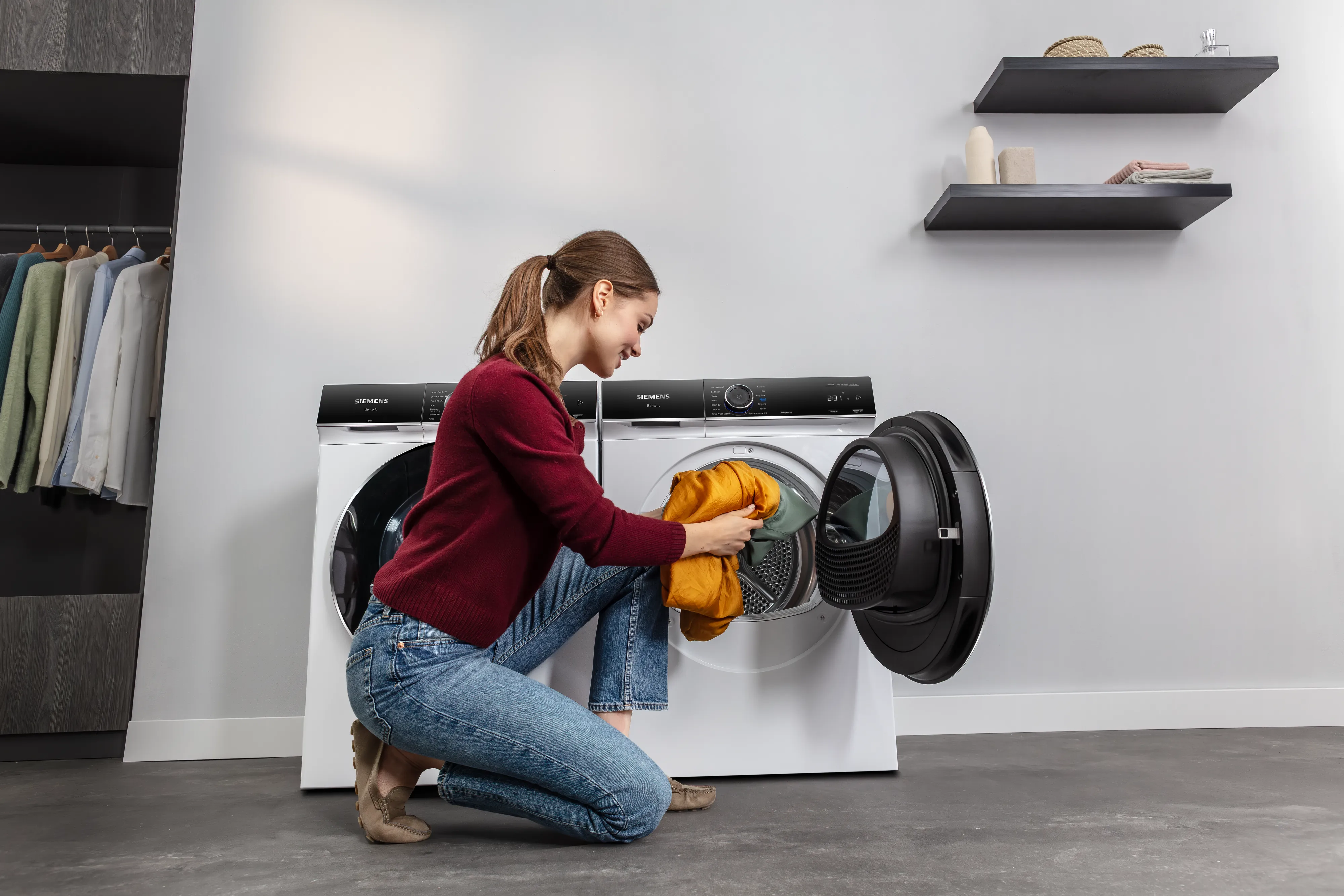 A woman wearing a burgundy sweater and jeans kneels in a modern laundry room, loading an orange garment into a white Siemens front-loading washing machine. An identical machine stands next to it, and there is an open wardrobe to the left.
