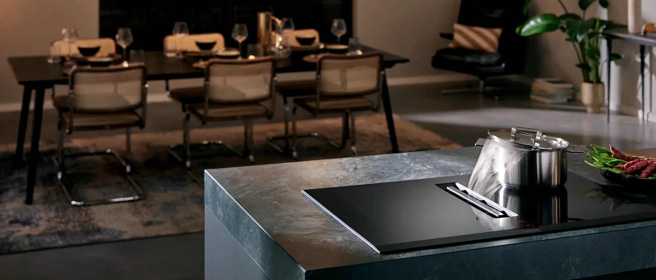 Modern kitchen with a sleek dark countertop, a steaming pot, and a dining table set for a meal in a stylish, cozy setting.