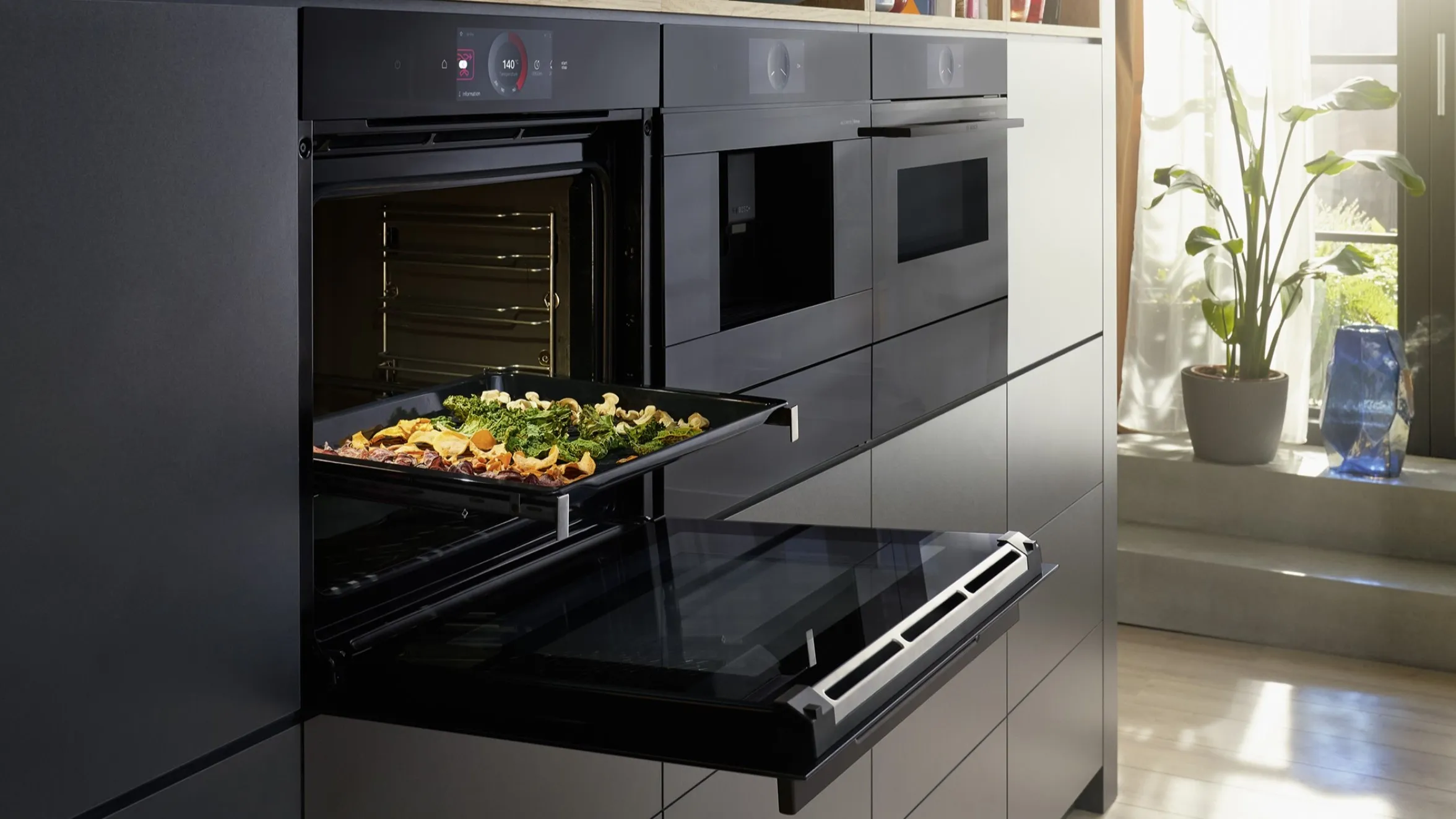 Een moderne zwarte wandoven met een bak geroosterde groenten gedeeltelijk uitgetrokken, in een stijlvolle keuken met daglicht 