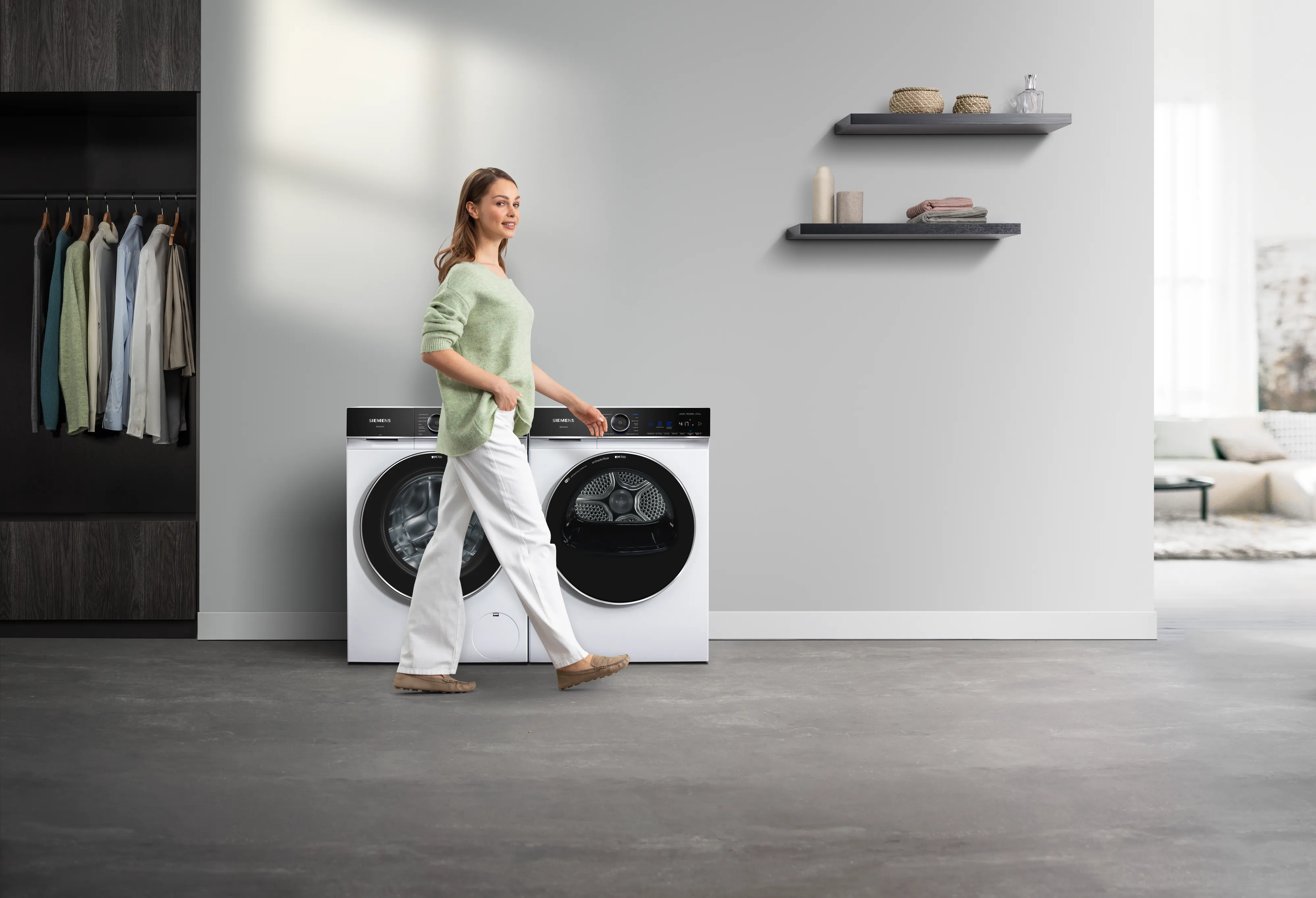 Een vrouw loopt langs een Siemens iQ700-wasmachine en -droger in een lichte wasruimte.