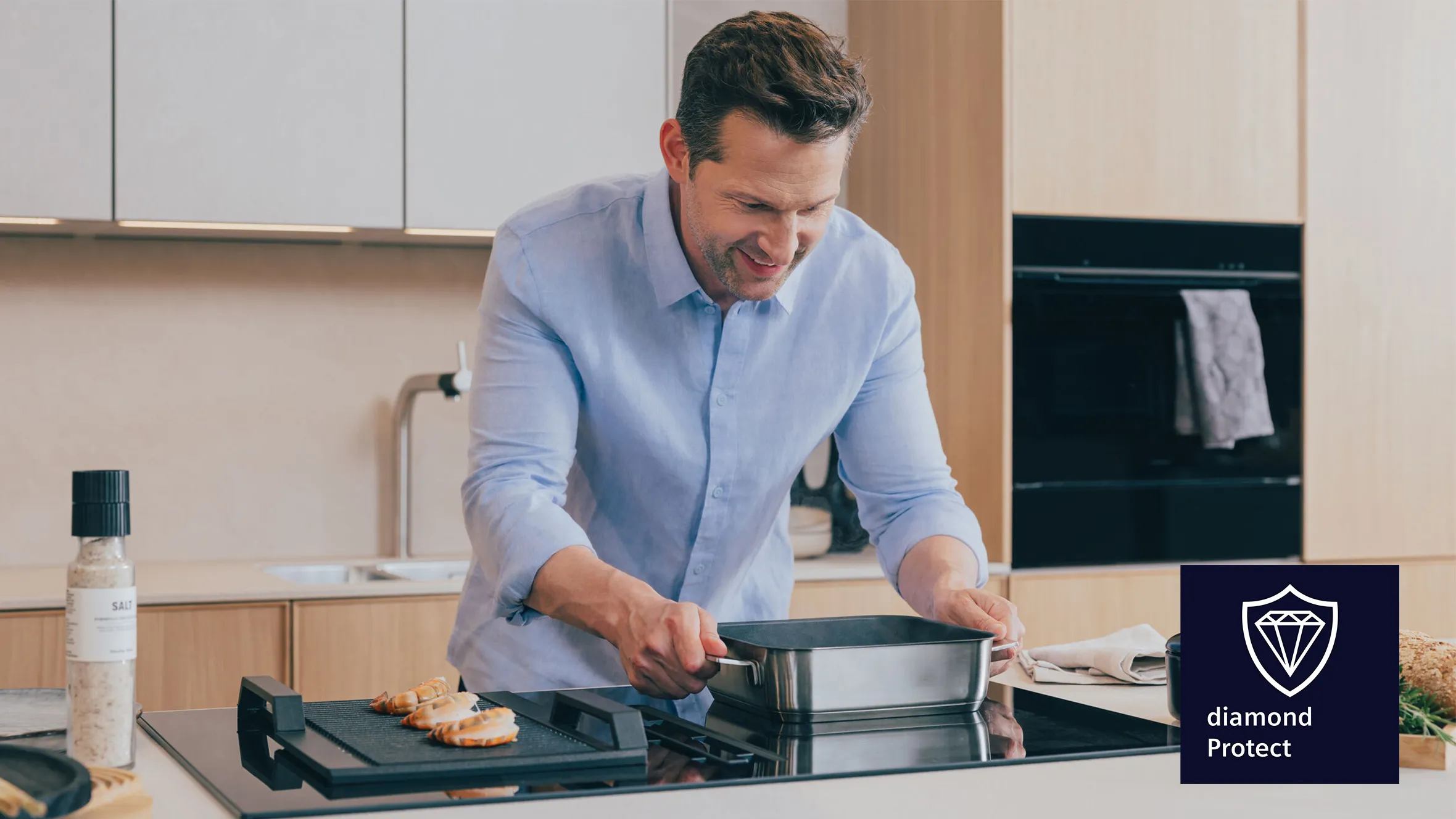 Un homme en chemise bleue en train de cuisiner avec une poêle sur une plaque de cuisson moderne, dans une cuisine lumineuse, où l'on aperçoit le logo « Diamond Protect ».