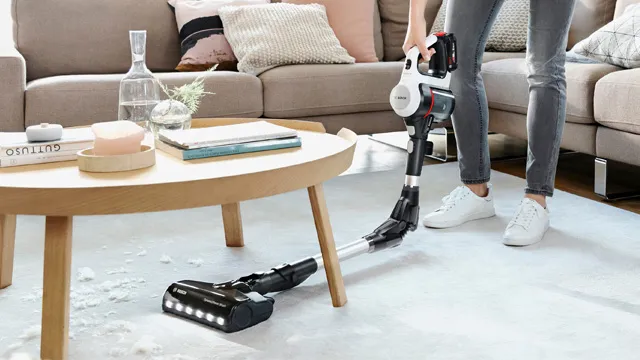 Eine Person benutzt einen kabellosen Staubsauger auf einem Wohnzimmerteppich und entfernt Schmutz unter einem Couchtisch. Im Hintergrund stilvolle, moderne Einrichtung.