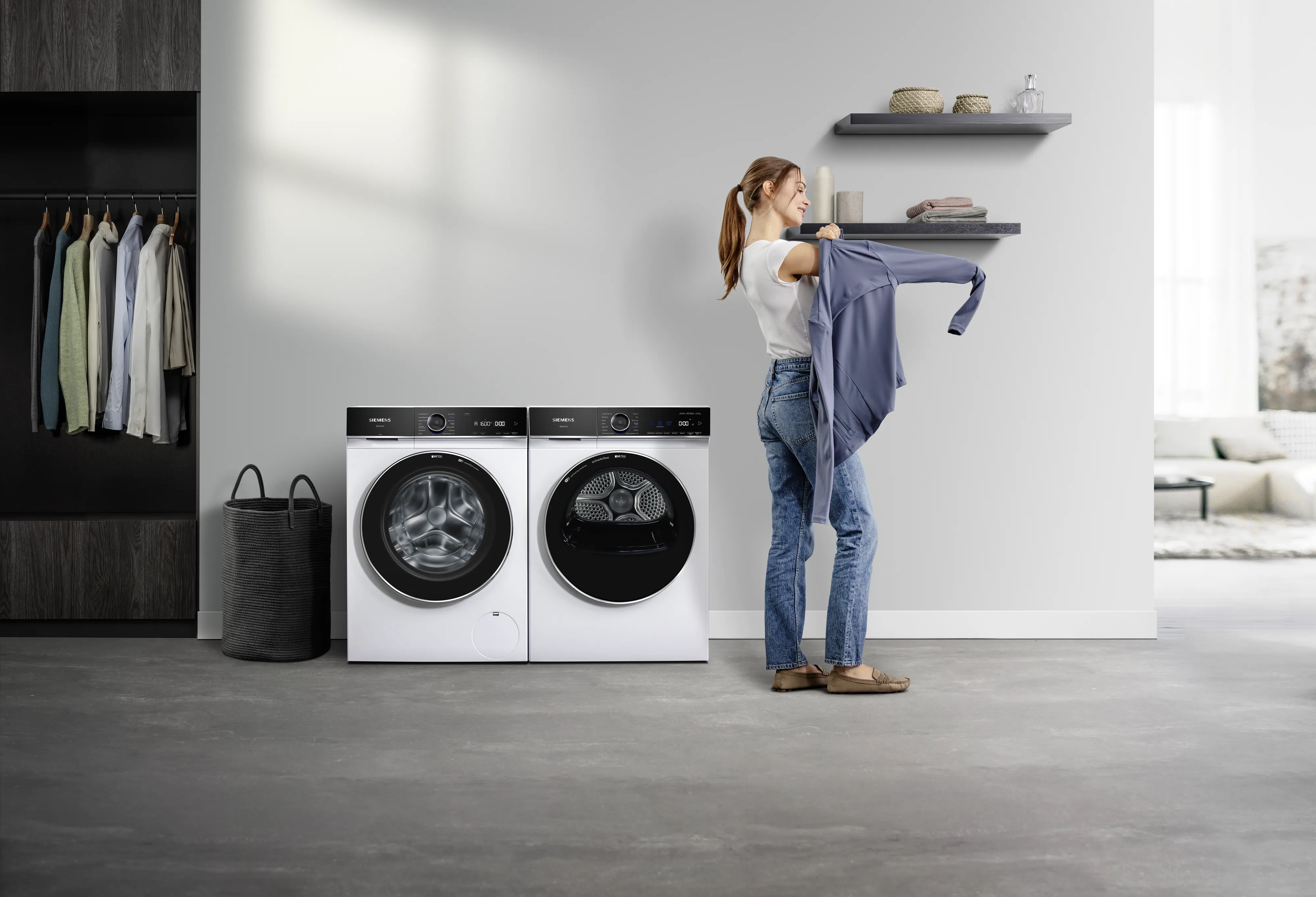 Eine Frau steht neben einem iQ700 Trockner und zieht sich eine Jacke an. Neben dem Trockner steht eine iQ700 Siemens Waschmaschine.