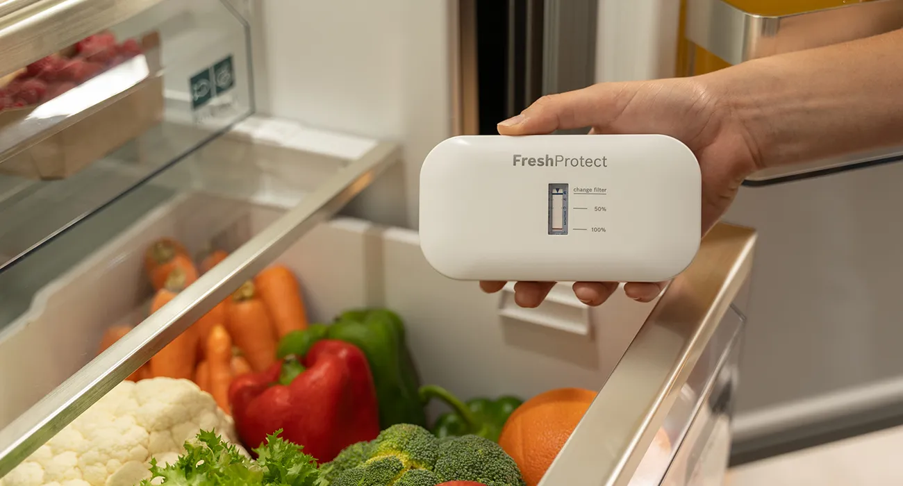 Main tenant un boîtier blanc marqué 'FreshProtect' devant un réfrigérateur ouvert rempli de légumes frais