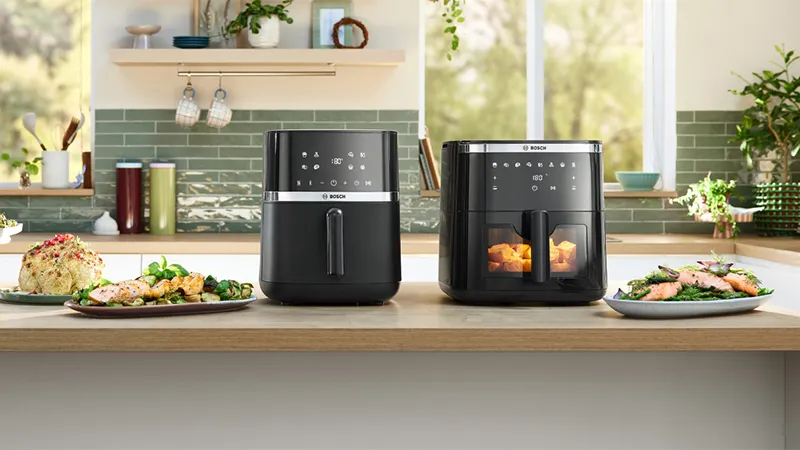 Zwei stilvolle schwarze Heißluftfritteusen stehen auf einer Küchentheke, umgeben von Tellern mit farbenfrohem, gesund zubereitetem Essen. Heller, luftiger Küchenhintergrund.