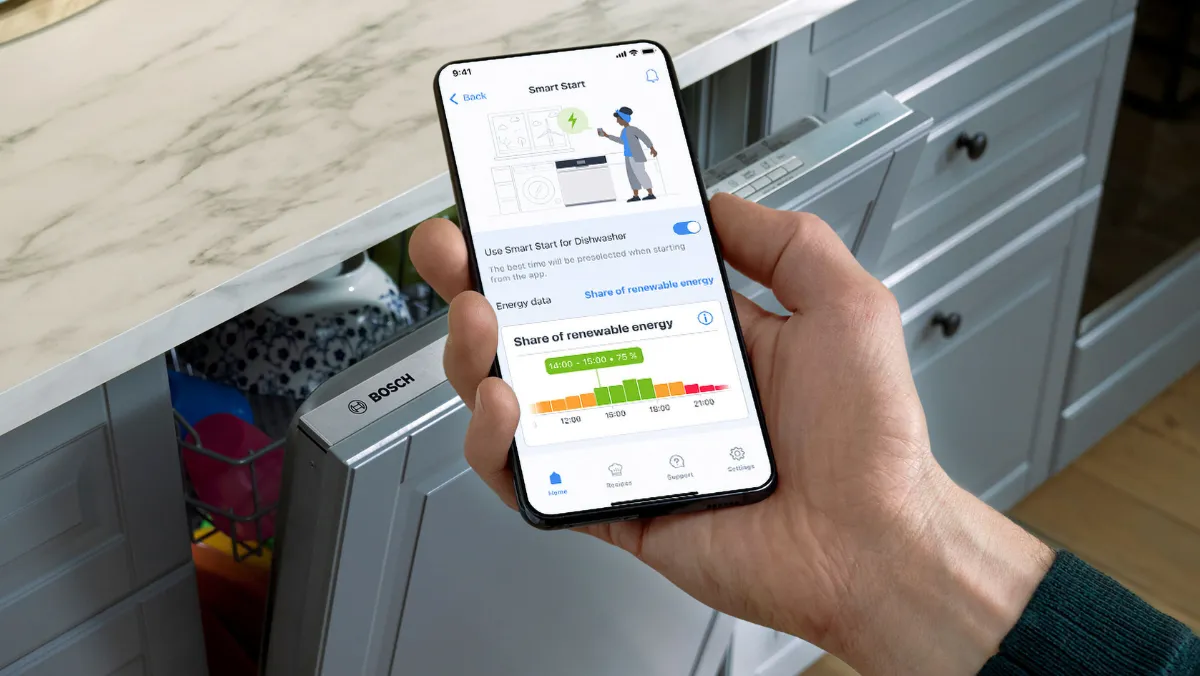 A person holding a smartphone displaying a smart appliance app, with a dishwasher partially open, on a countertop with a marble surface.