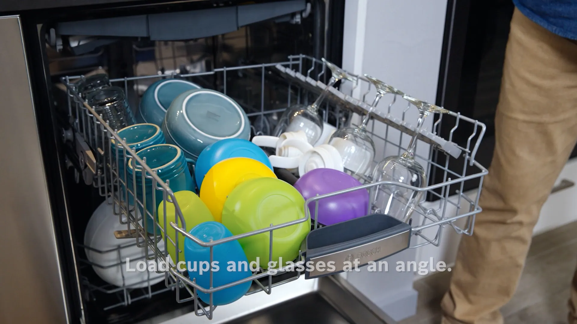Open Bosch dishwasher loaded with colorful cups and bowls at an angle. Text reads: Load cups and glasses.