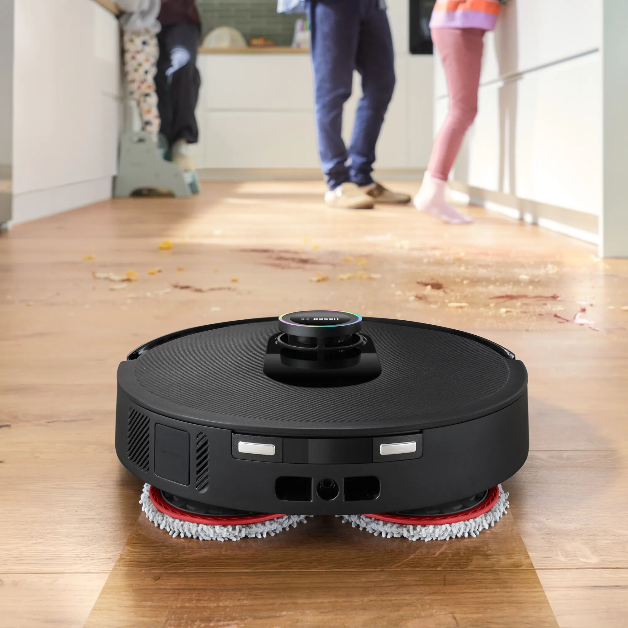 A black Bosch robot vacuum and mop cleans crumbs and spills on a wooden kitchen floor while a family stands in the background.