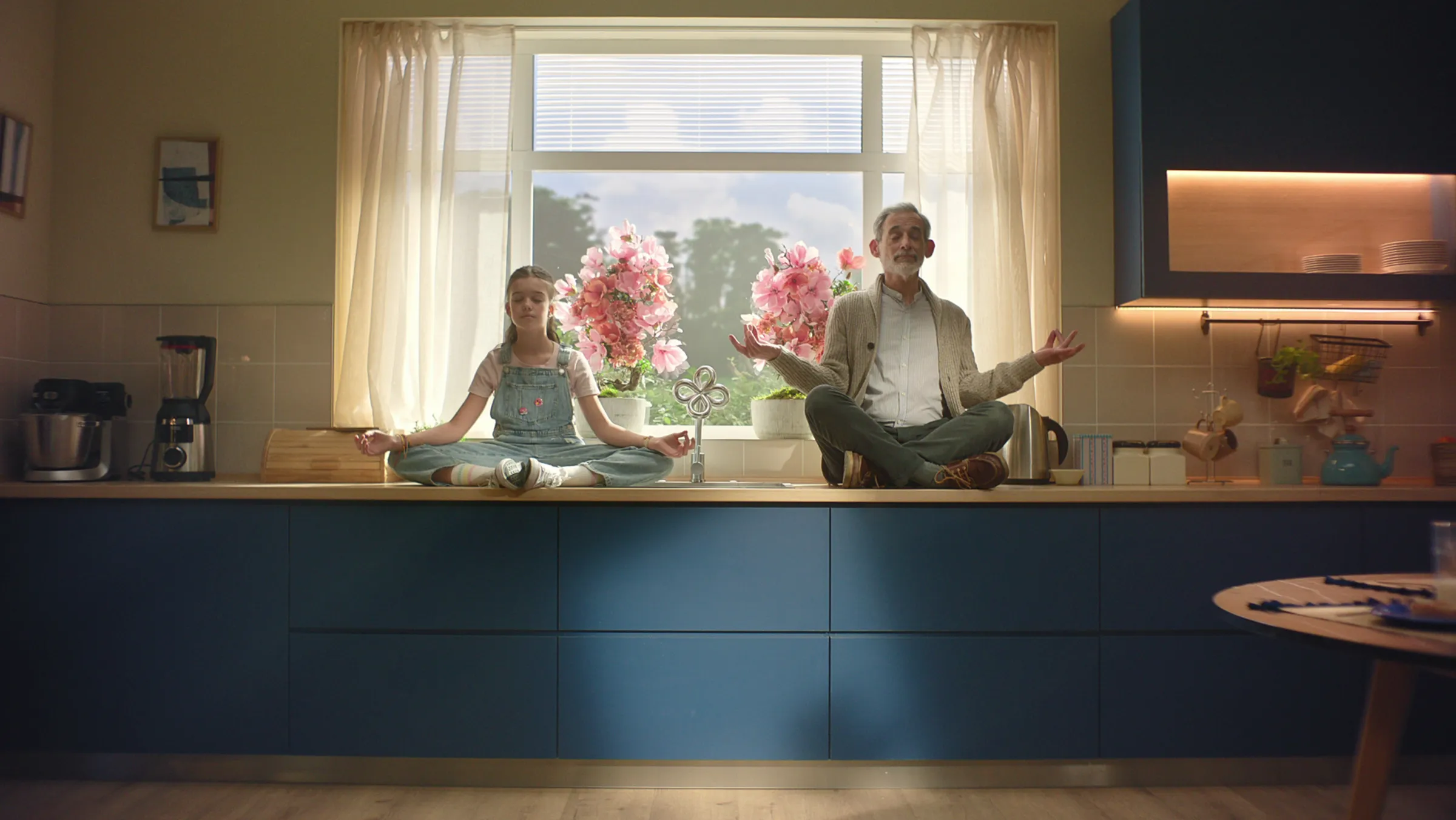 A man and a girl sitting cross-legged on a kitchen counter in front of a window with curtains and flowers, both meditating with eyes closed