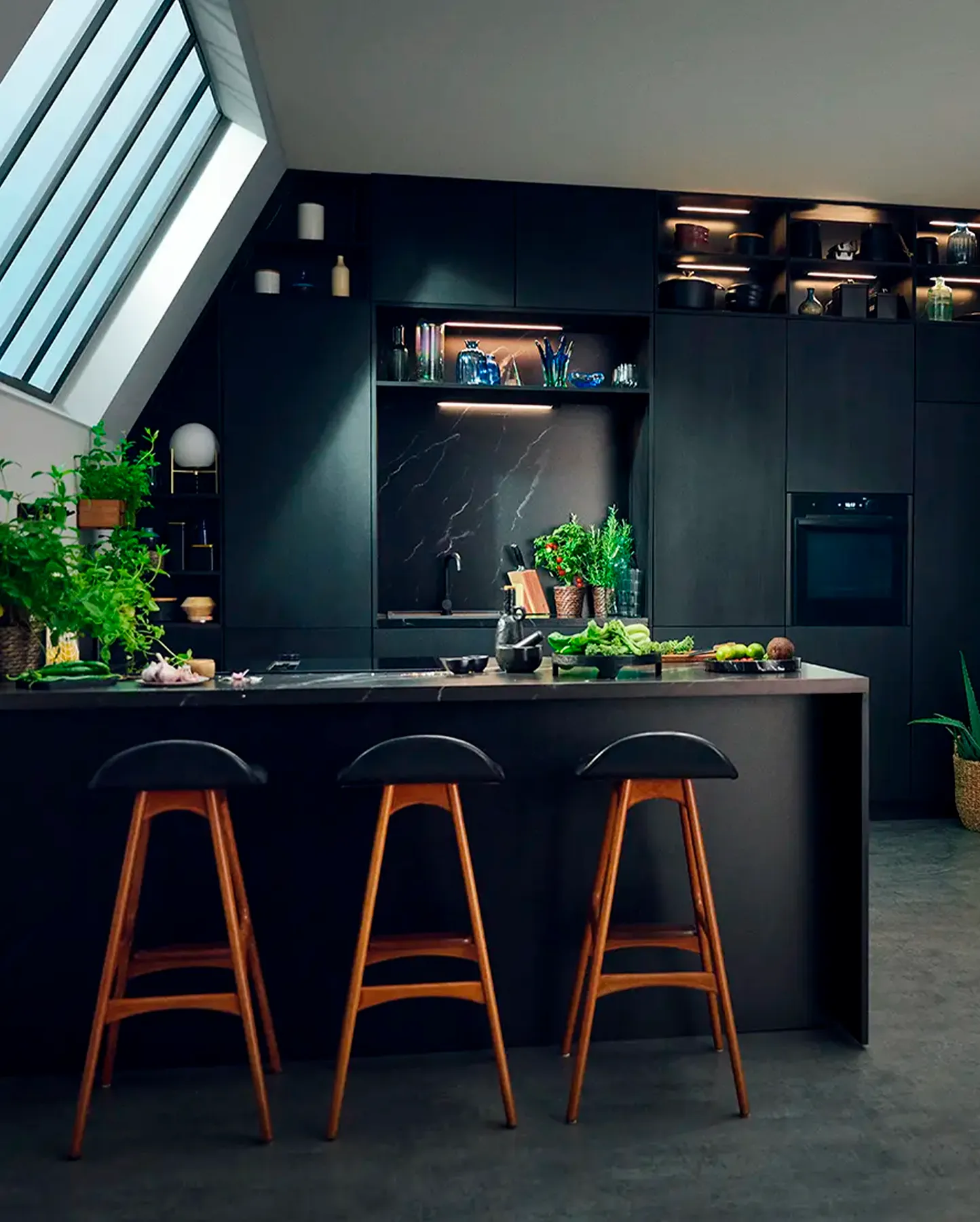 „O bucătărie cu suprafețe închise la culoare și o insulă de bucătărie înaltă, iar în spate se află un cuptor încorporat.