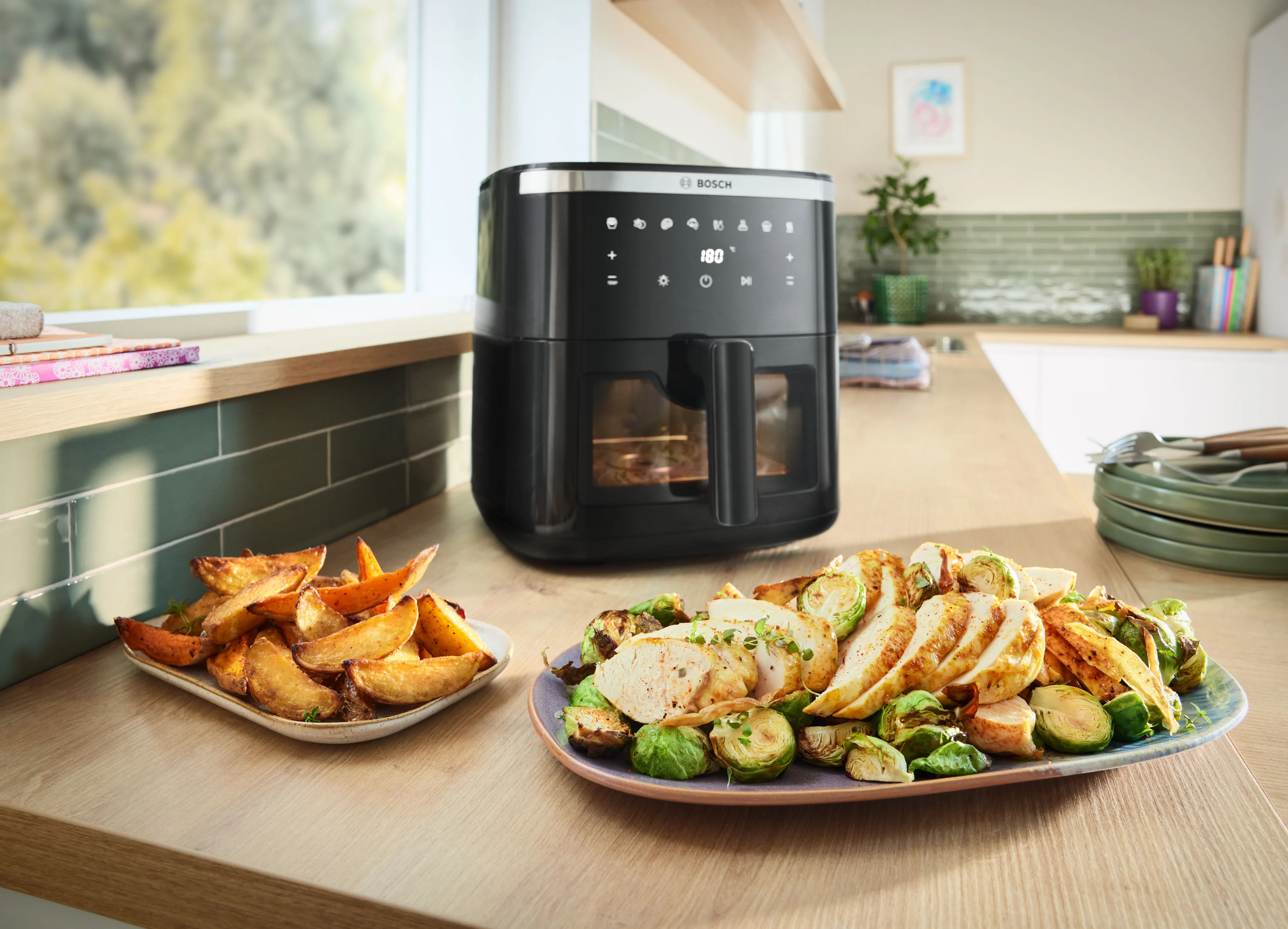 A plate with tender salmon fillets, crisp asparagus and golden roasted potatoes sits in front of a Bosch Air Fryer on a kitchen countertop, showcasing the appliance’s energy-efficient and fast cooking capabilities.
