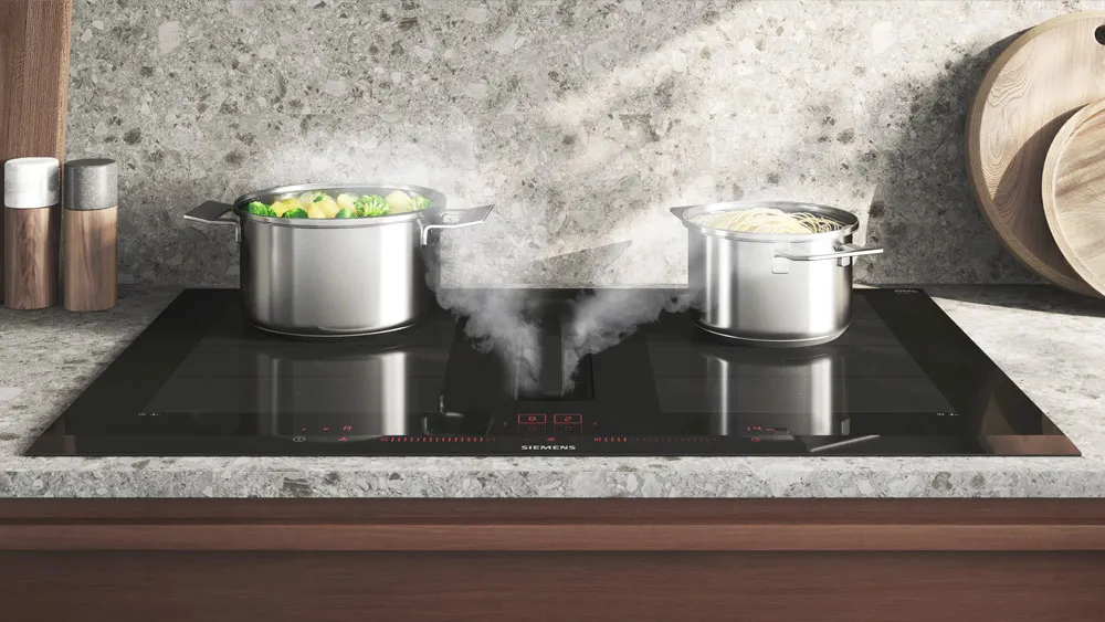 Piano cottura a induzione nero con due pentole in acciaio inox da cui esce vapore, sullo sfondo parete in pietra chiara e due contenitori cilindrici in legno.
