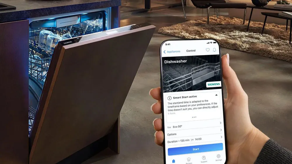 Lavastoviglie con sportello aperto in cucina moderna e mano che tiene uno smartphone con app di controllo lavastoviglie visibile