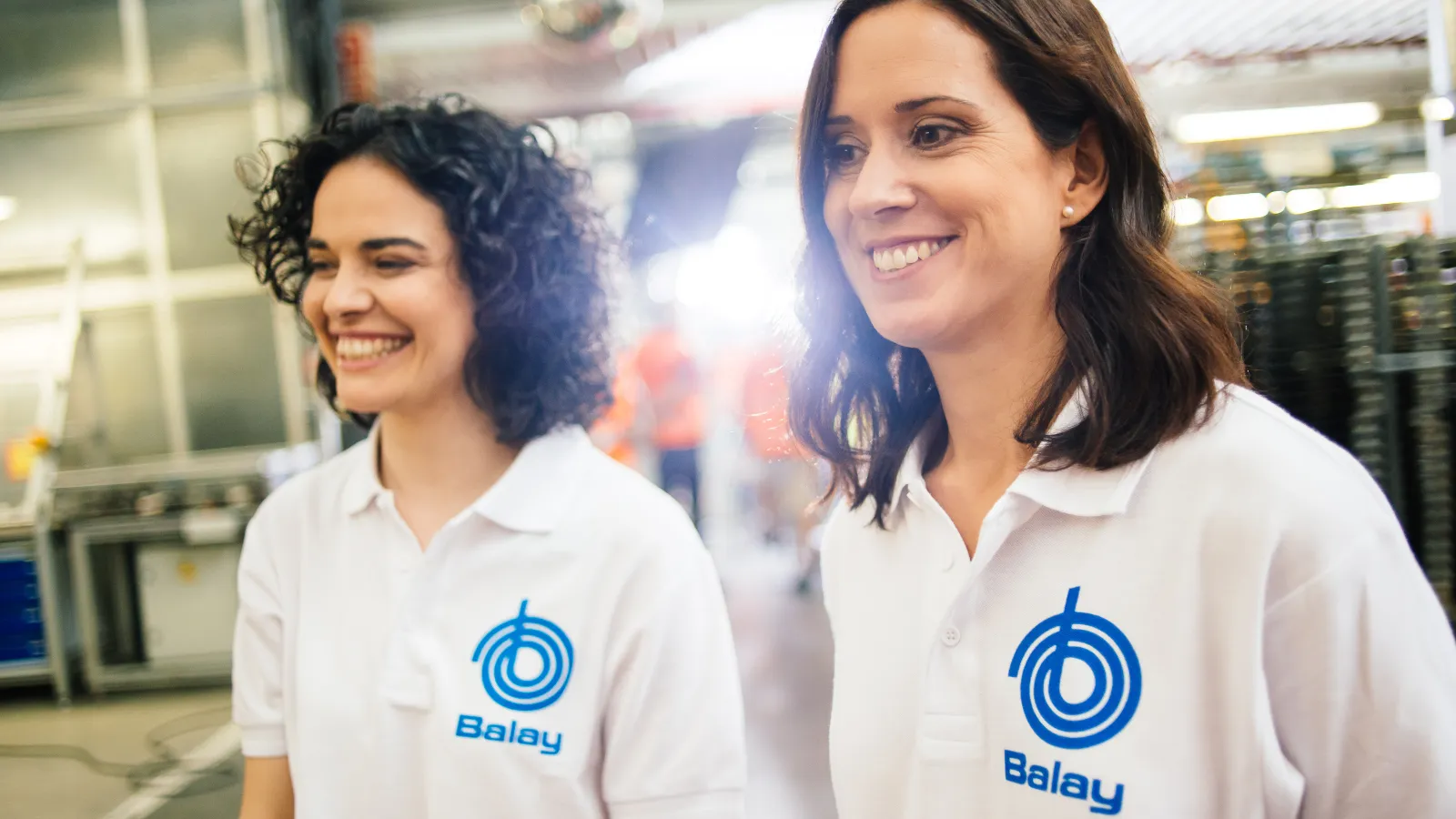Dos personas con polos blancos con el logotipo "Balay" de pie en un entorno industrial o de fábrica.