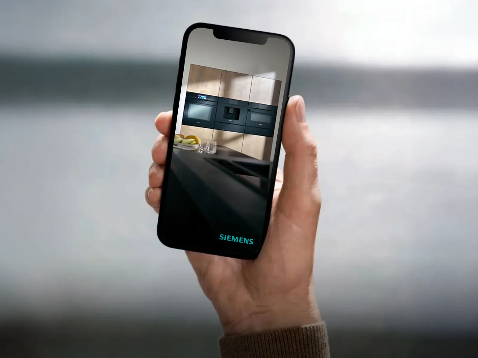 A person holds a smartphone displaying a modern Siemens kitchen with sleek appliances against a light wood backdrop.