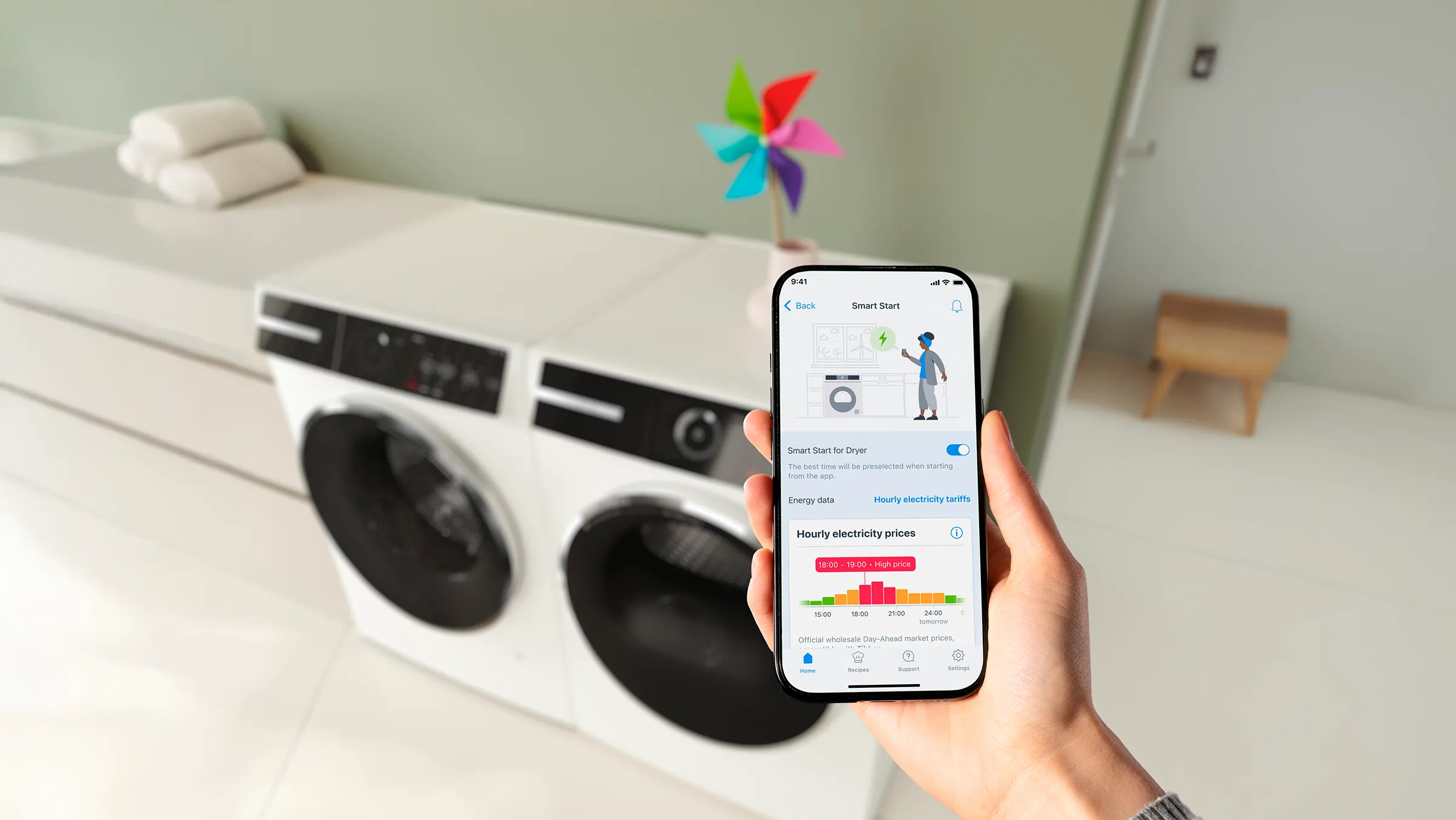 Eine Hand hält ein Smartphone mit der aktivierten Smart-Start-Funktion in der Home Connect App vor einer Bosch Waschmaschine und einem Trockner.