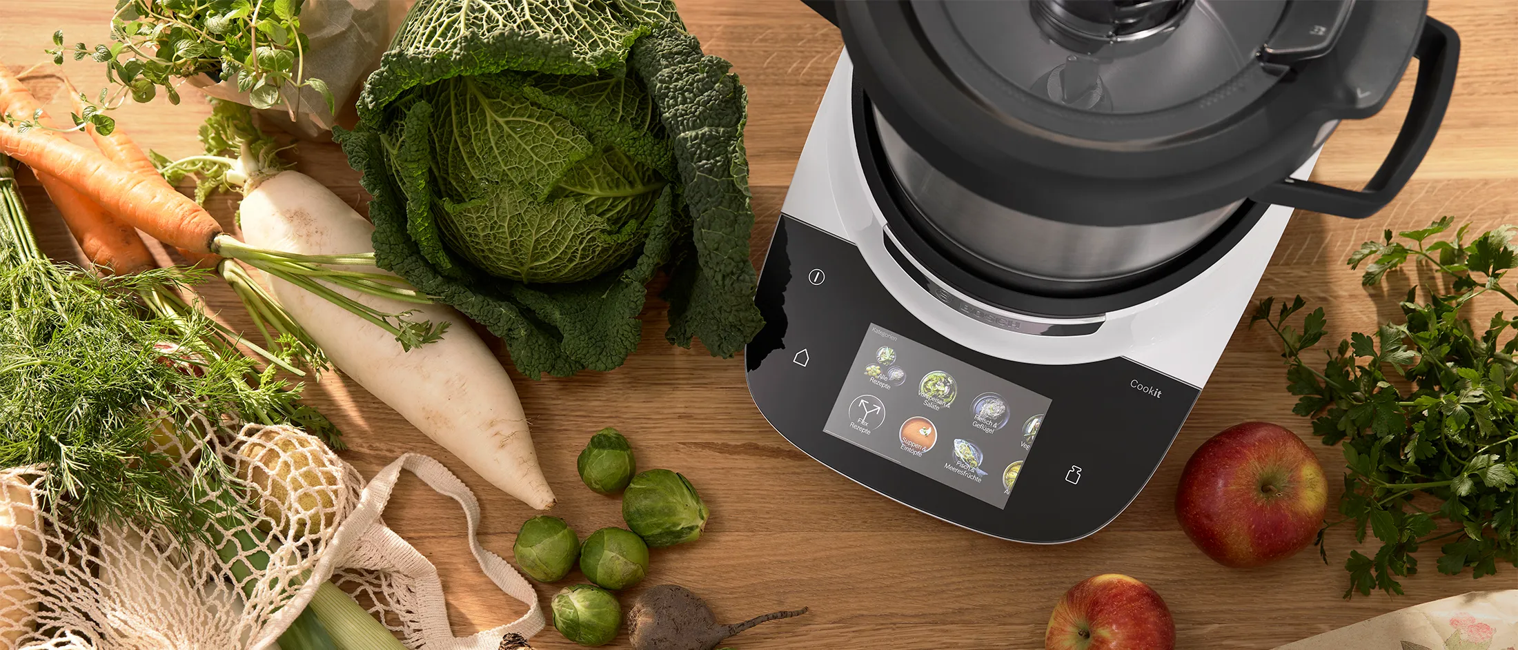 Bosch food processor with touch display surrounded by various fresh vegetables on a table.