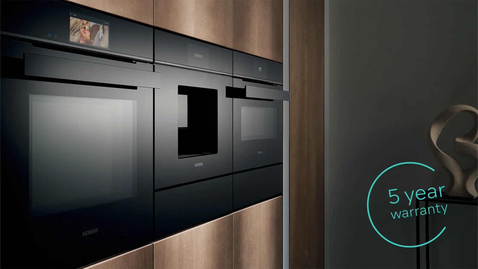 A sleek, modern kitchen featuring black built-in ovens with a 5-year warranty stated in a circular graphic. Warm wooden paneling surrounds them.
