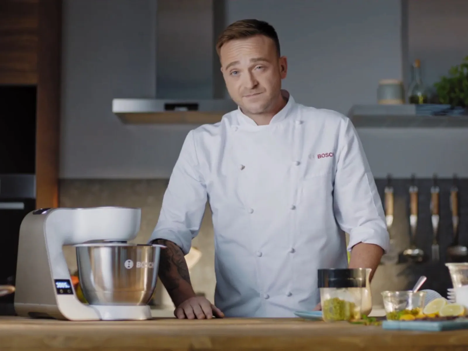 Mateusz Gessler kucharz w białym uniformie stoi w nowoczesnej kuchni, na blacie robot planetarny Bosch.