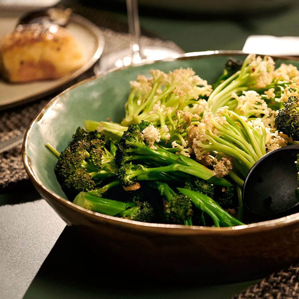 A bowl of sautéed broccoli and cauliflower florets, garnished and served alongside a small, golden-brown bread roll.