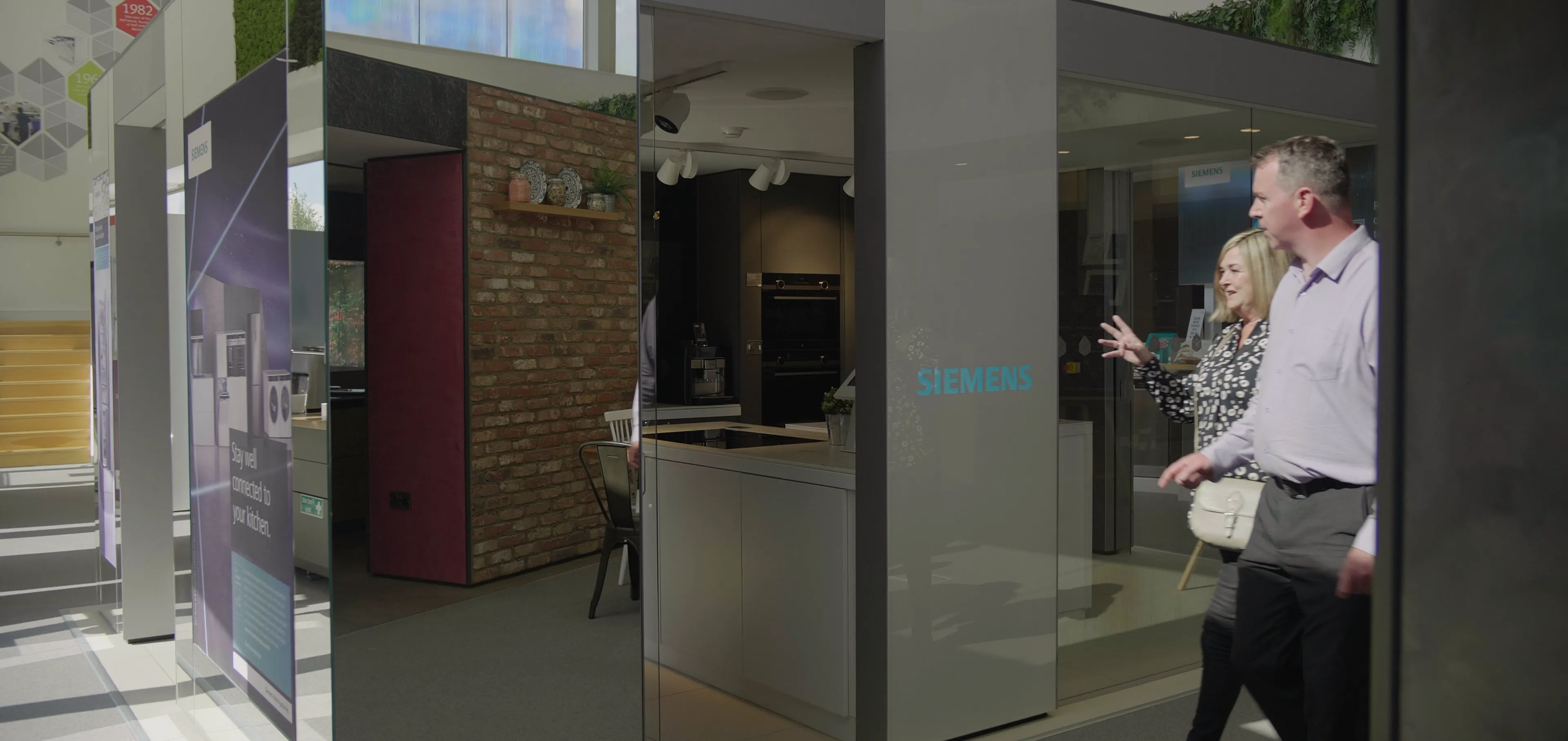 A man and a woman are walking into a modern Siemens appliance showroom. The woman gestures enthusiastically towards a kitchen display area that is visible through a glass entrance, while the man looks on. The showroom features various displays and informational banners.