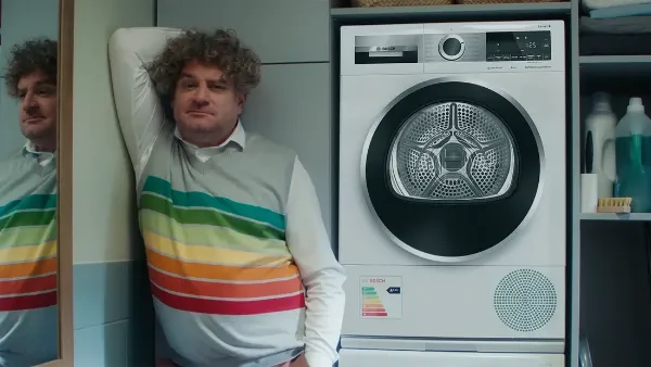 Een persoon leunt tegen een spiegel, gekleed in een kleurrijke gestreepte trui, naast een moderne wasmachine in een lichte wasruimte.