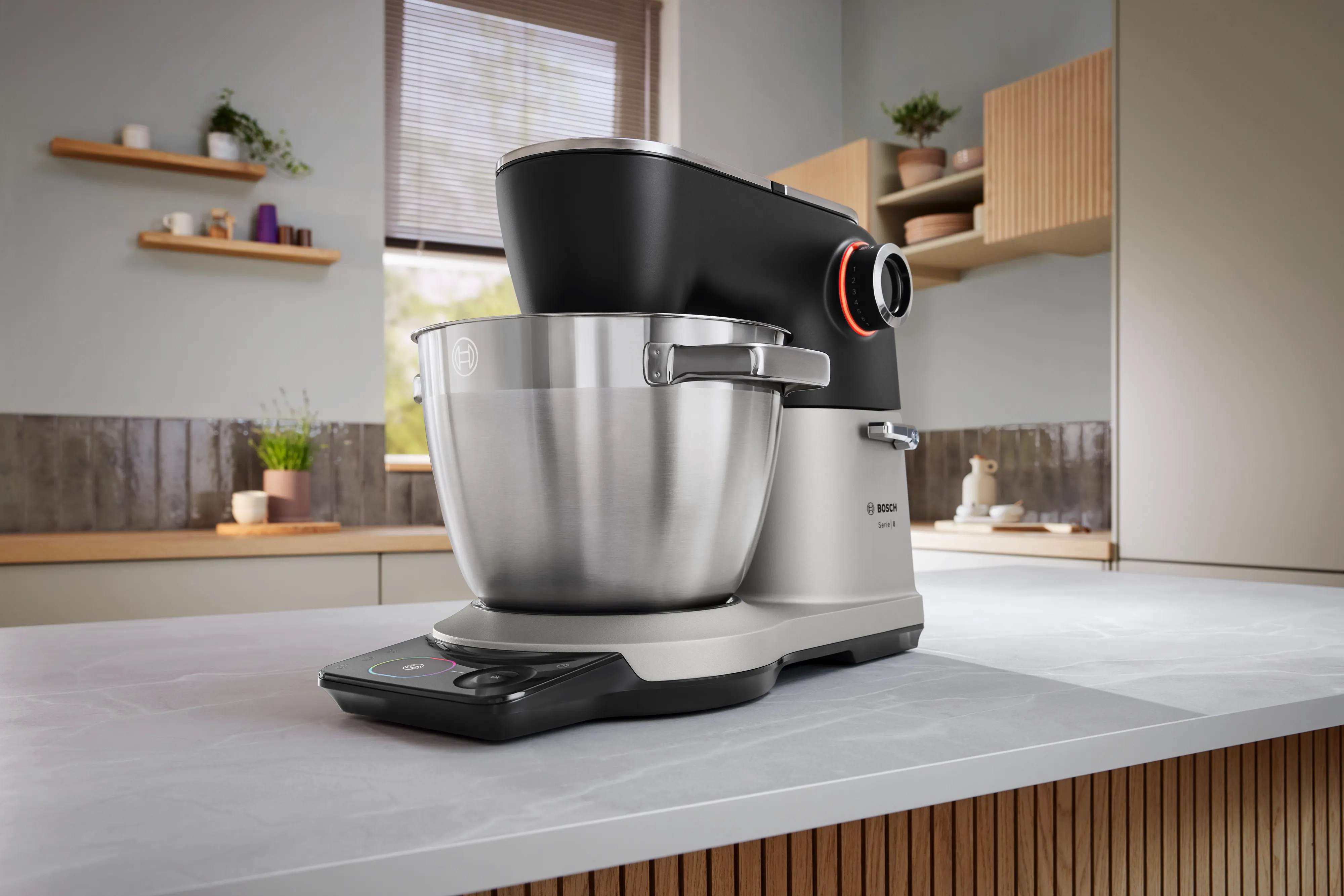Un elegante miscelatore da cucina Bosch con una ciotola in acciaio inossidabile, sullo sfondo di una cucina moderna con accenti e piante in legno.