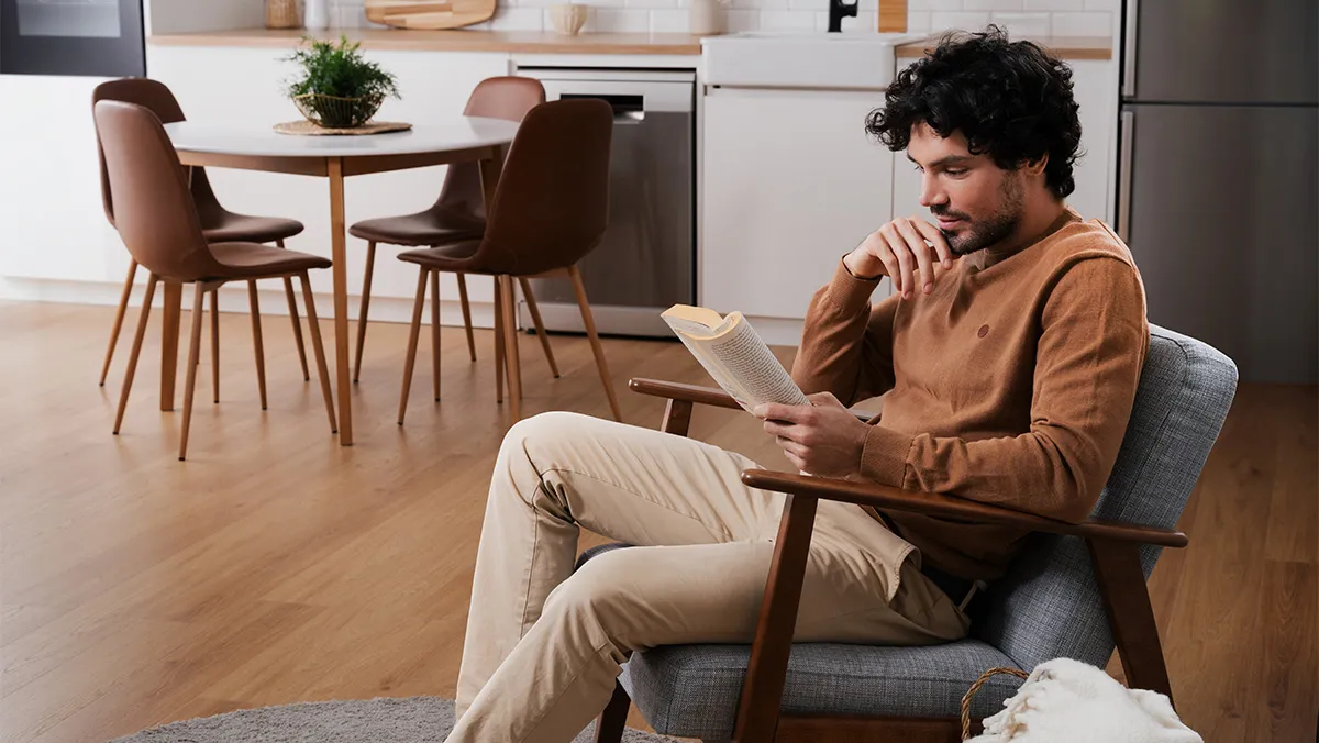 Homem sentado num cadeirão a ler um livro numa moderna kitchenette com piso de madeira e cadeiras castanhas.