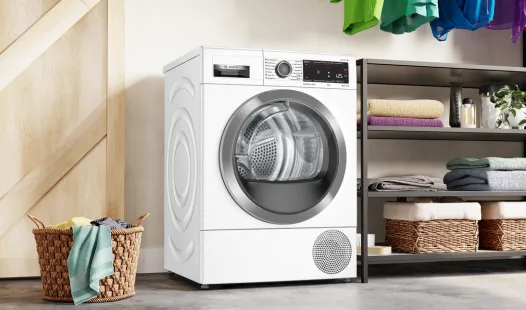 A white Bosch heat pump dryer stands next to a wooden door and a metal shelving unit with folded laundry and baskets.