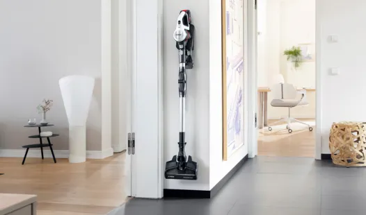 A Bosch Unlimited 7 cordless stick vacuum cleaner is mounted on a white wall in a modern home, with a light-colored room and furniture visible through a doorway on the left