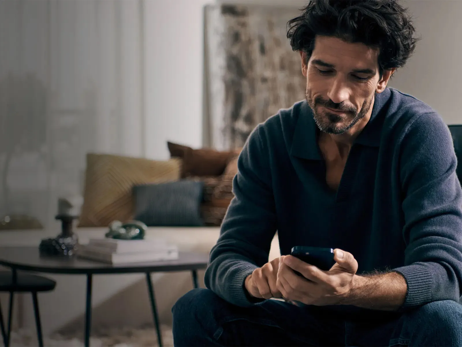 Man sitting on a couch using a smartphone, with pillows and a coffee table in the background.