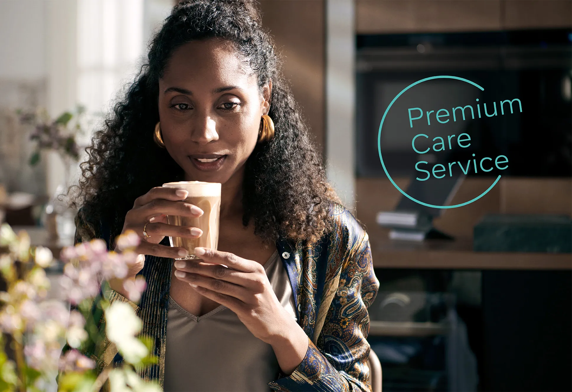 Kvinde, der nyder en kaffe i stuen med blomster og Premium Care Service logo.